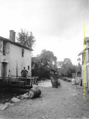1 vue - Rue de village : un homme sur une passerelle de bois devant sa maison et des tonneaux dans la rue. - [entre 1900 et 1920]. - Photographie (ouvre la visionneuse)